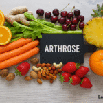 Photo d’une composition colorée d’aliments anti-inflammatoires – carottes, fraises, cerises, oranges, citron, ananas, céleri, gingembre et noix – disposés sur une table en bois claire autour d’une ardoise noire marquée « Arthrose ». Image réaliste et lumineuse, illustrant une alimentation saine pour soulager les douleurs articulaires.